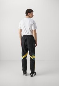 Pantalones deportivos negros con rayas en forma de cheurón verdes y amarillas, cinturilla elástica y una tela ligera, combinados con una camiseta blanca y zapatos negros.