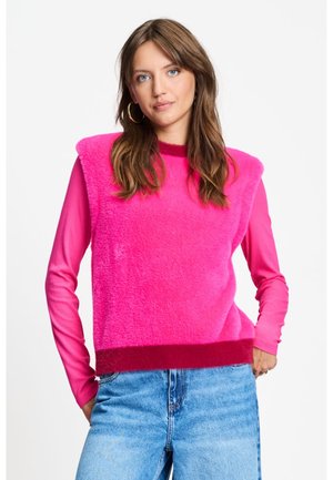 Vrouw met lang bruin haar, die een felroze mouwloze pluizige top draagt over een roze longsleeve en lichtblauwe spijkerbroek.