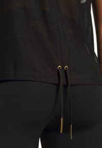 Haut en tissu maille noire avec cordons noirs ornés d'embouts métalliques dorés, porté sur un pantalon noir ajusté, gros plan sur la partie inférieure du dos.