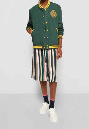 Veste bomber verte avec accents jaunes et emblème, associée à une robe rayée aux couleurs multiples, des chaussures blanches et des chaussettes noires.