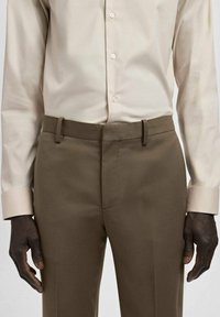 Hellbeige Hemd mit figurbetontem Schnitt und Knopfleiste vorne, kombiniert mit dunkelbraunen, maßgeschneiderten Hosen mit Gürtelschlaufen.