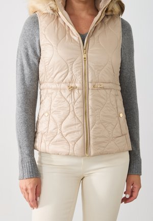 Kvinde iført beige quiltet vest med pelsforet hætte, guld lynlås og snøre over en grå langærmet sweater og cremefarvede bukser.