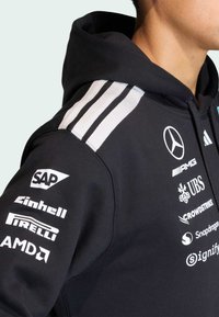 Sweat à capuche noir avec cordon de serrage, comportant trois bandes blanches sur l'épaule et plusieurs logos blancs imprimés sur la manche et la poitrine.