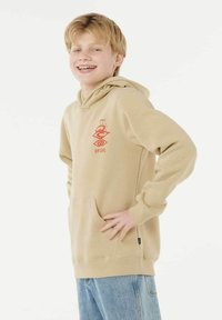 Beige hoodie met een voorzak en een rood grafisch logo. Lange mouwen met geribde manchetten, gemaakt van zachte stof, gedragen over blauwe jeans.