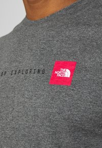 Záběr zblízka na šedou košili s bílým logem "THE NORTH FACE" na červeném čtverci a částečným černým textem "STOP EXPLORING."