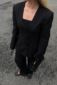 Blazer nero con scollatura quadrata, grandi bottoni e spalle strutturate; abbinato a pantaloni coordinati e scarpe nere texturizzate; tiene una borsa nera.