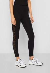 Leggings noirs avec un panneau latéral en dentelle texturée, dotés d'un design ajusté et d'un tissu lisse et extensible. Portés avec des chaussures de sport blanches.
