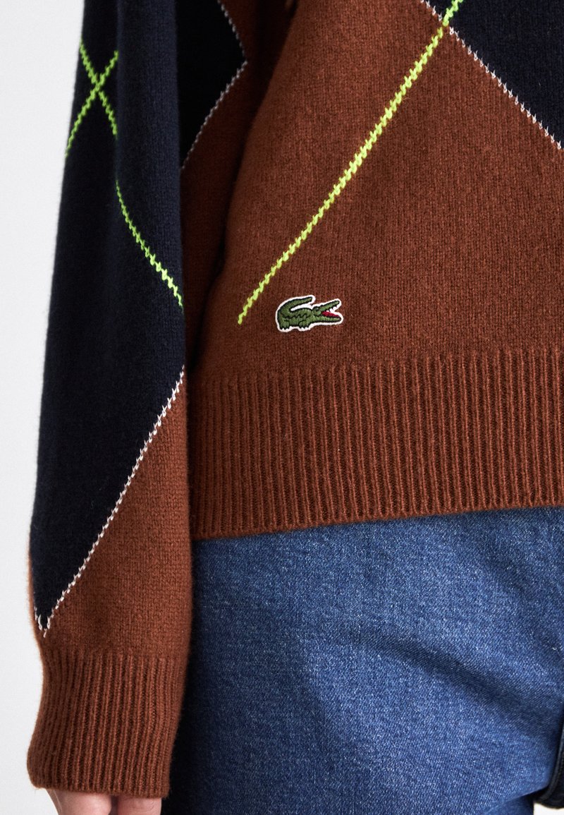 Pull en tricot avec un corps marron orné de motifs en losange bleus et verts, des poignets côtelés et un petit logo de crocodile en accent.