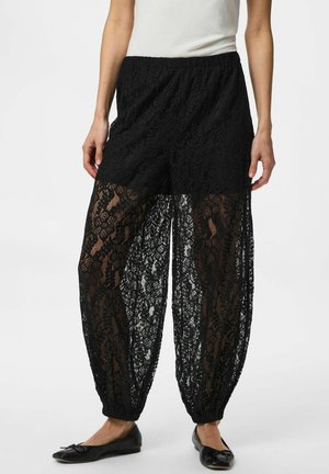 Nő fekete csipkés nadrágot visel elasztikus mandzsettákkal, fekete rövidnadrág felett, fekete balettcipővel és fehér felsővel párosítva.