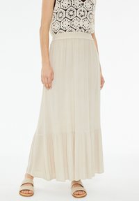 Falda maxi beige, fluida, con dobladillo en capas, tejido ligero y cinturilla elástica. Llevada con sandalias suaves y en tonos neutros.