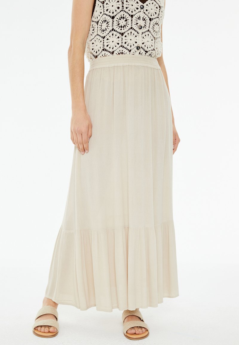 Falda maxi beige, fluida, con dobladillo en capas, tejido ligero y cinturilla elástica. Llevada con sandalias suaves y en tonos neutros.