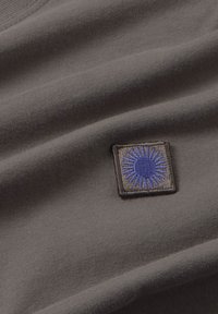 Grijze katoenen stof met een textuur, voorzien van een vierkant blauw geborduurd stukje met zonnestraaldesign op een bruine achtergrond.