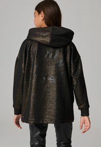 Sudadera negra y dorada con purpurina, con capucha, de corte oversized y un diseño de texto llamativo en la espalda. Textura suave con puños acanalados.