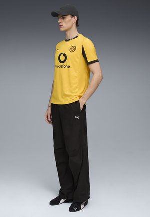 Geel short-sleeve voetbalshirt met zwarte accenten, voorzien van een cirkelvormig Vodafone-logo en een clubembleem, gecombineerd met zwarte trainingsbroeken en schoenen.