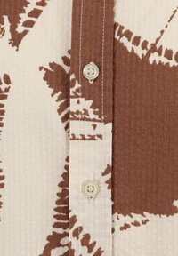 Camicia a maniche corte marrone e crema, caratterizzata da un tessuto testurizzato, chiusura con bottoni e un motivo botanico. Cuciture evidenti lungo i bordi.