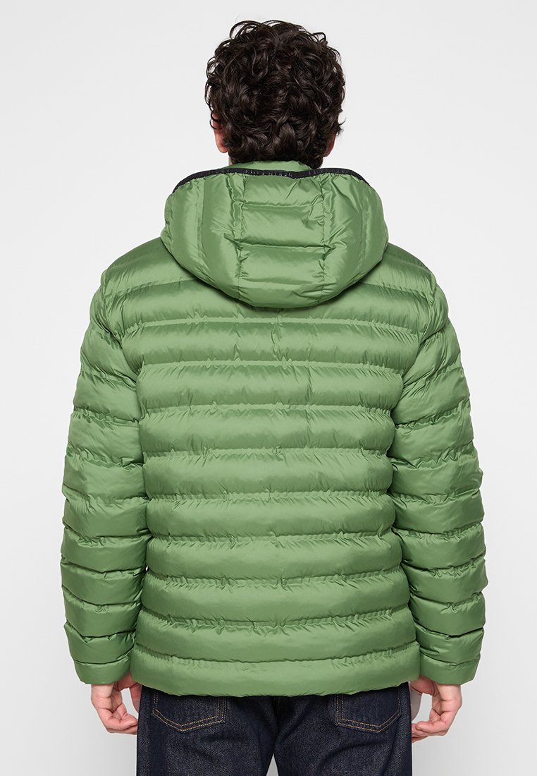 Persona vista di spalle che indossa un giubbotto imbottito verde con cappuccio e jeans scuri su uno sfondo semplice.
