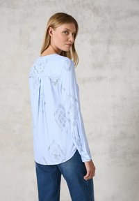 Hellblaues Langarmshirt mit ausgeschnittenen Diamantmustern, geschichtetem Rücken, weichem Stoff und lockerem Schnitt, kombiniert mit dunkler Jeans.