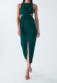 Vrouw in een mouwloze donkergroene midi-jurk met zijsneetjes en een split aan de voorkant, gecombineerd met gouden bandjeshakschoenen en een gouden armband.