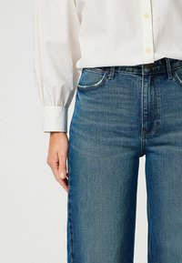 Jeans de mezclilla azul de cintura alta y pernera ancha con cinco bolsillos y un ligero desvanecimiento. Combinados con una camisa blanca de manga larga con puños abotonados.