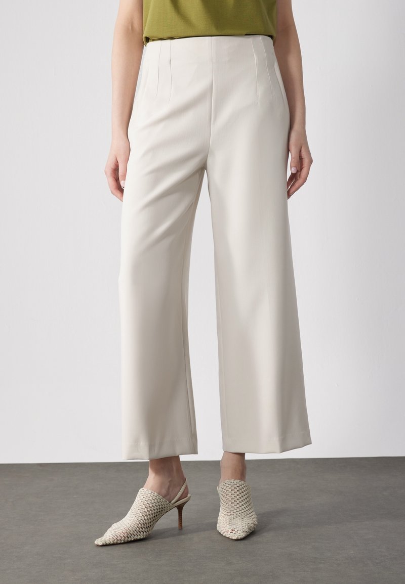 Personne portant un pantalon crème taille haute à jambes larges et des mules tissées beiges à talons, debout sur un sol gris contre un mur blanc.