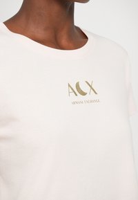 Svetlo roza bombažna majica z zlatim folijskim logotipom "ACX" in besedilom "ARMANI EXCHANGE", z okroglo izrezano ovratnikom in kratkimi rokavi.
