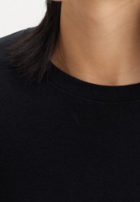 Svart tröja med ribbad nacke. Korta svarta hår, slät hudton. Enkel, minimal design, inga synliga mönster.