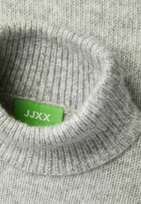 Svijetlo sivi pleteni turtleneck pulover s rebrastom teksturom. Ima zeleni etiketu s bijelim tekstom "JJXX" koja je vidljiva unutar ovratnika.