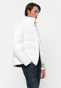 Man in een witte pufferjack met hoge kraag, donkere jeans en een zwart shirt, staande in profiel met de handen in de zakken tegen een witte achtergrond.