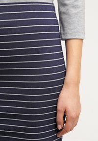 Pruhovaná námořnická sukně se světle šedými vodorovnými pruhy. Tkanina se zdá být pružná; nošena s šedým dlouhým rukávem. Ruka je opřená o sukni.