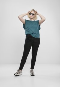 Teal överstor t-shirt knuten i midjan, svarta slim fit jeans och grå sportskor med svarta accenter. Tillbehören inkluderar svarta solglasögon.