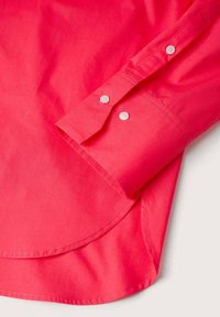 Poignet de chemise rose vif à manches longues avec boutons blancs, ourlet incurvé visible et coutures nettes sur un fond clair.