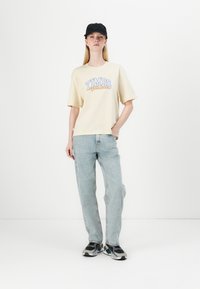 Šviesiai geltona per didelė t-shirt su tekstu "TIMBS Authentic", derinama su plačiomis šviesiai mėlynomis džinsomis ir juodais sportbačiais.