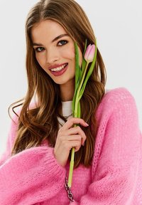 Różowy puchaty sweter z szerokimi rękawami, trzymany przez osobę, która się uśmiecha i trzyma różową tulipana. Jasne tło podkreśla kolory.