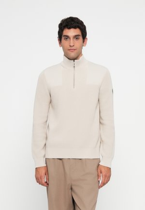 Man with dark curly hair wearing a cream half-zip sweater and beige pants, standing against a plain white background.