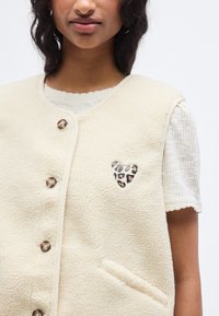 Μπεζ γιλέκο από φλις με στ ρογγυλεμένο ντεκολτέ, κουμπιά ως κούμπωμα και λεπτομέρεια καρδιάς με λεοπαρ μοτίβο στο στήθος. Διαθέτει πλαϊνές τσέπες.