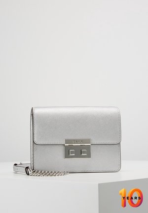 Sac bandoulière rectangulaire argenté avec surface texturée, fermoir en métal et chaîne, posé sur un piédestal blanc sur fond gris clair.