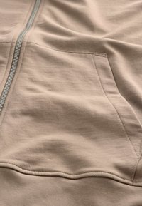 Beige zip-up beklædning med en glat tekstur og en frontlomme; har en metallisk lynlås og rene syningsdetaljer.