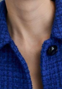 Modrý tvídový kabát s texturovanou látkou, zdobený kulatým černým knoflíkem a límcem, který rámuje krk.