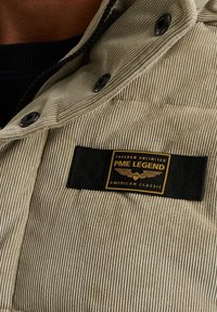 Vaalea corduroy-takki mustilla neppareilla, jossa on näkyvä kultainen ja musta etiketti, jossa lukee "PME Legend American Classic."