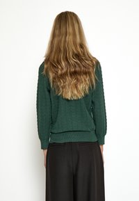 Suéter verde de punto con un patrón de olas texturizado, puños y dobladillo acanalados, combinado con pantalones de pierna ancha negros. Vista desde la parte posterior.