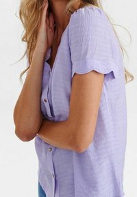 Donna che indossa una blusa a maniche corte color lavanda con bordi scallop, che appoggia una mano sul collo e l'altra sulla vita.