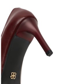 Bordeaux hoge hak schoen met een soepel leren bovenwerk, taps toelopende hak, zwarte zool en gouden logo accent aan de zijkant van de hak.