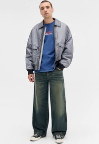 Šviesiai pilka bombonešio striukė su užtrauktuku, mėlynas grafikos marškinėlis, platūs tamsiai žali džinsai ir juodi sportbačiai, sukuriantys sluoksniuotą laisvalaikio aprangą.