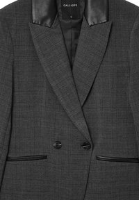 Blazer à carreaux gris avec revers en cuir noir, présentant un design croisé, deux boutons et des poches latérales avec des accents noirs.