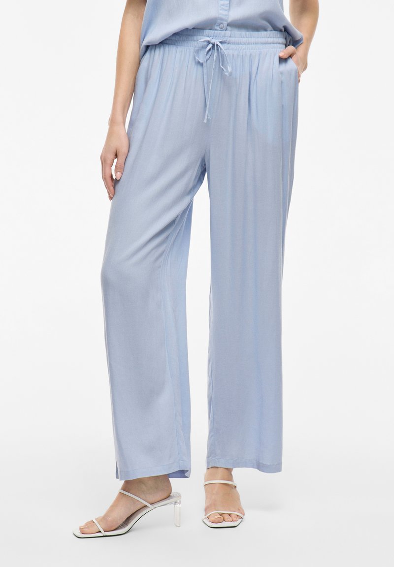 Gai zili pelēku platā kāju bikses, kas izgatavotas no mīksta, gluda auduma ar elastīgu jostu un auklu. Saskaņotas ar caurspīdīgām papēža sandālēm.