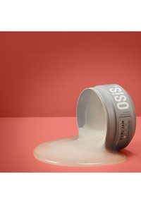 Grå cylindrisk behållare som tippar över, med en krämig produkt som rinner ut. Bakgrunden är en enfärgad korallfärg.