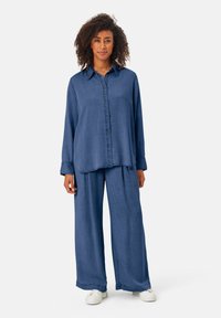 Tenue en denim composée d'une chemise à manches longues et boutons et d'un pantalon large, tous deux dans une teinte bleue uniforme, avec une texture lisse et une coupe décontractée.