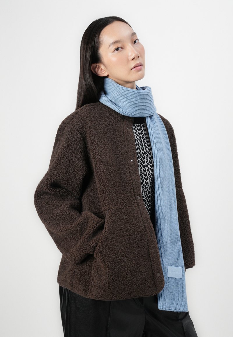 Braune Fleecejacke mit Knopfverschluss, ausgestattet mit einer strukturierten Oberfläche, kombiniert mit einem hellblauen gerippten Schal. Beide Teile sind über gemusterter Kleidung geschichtet.