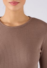 Maglietta a maniche lunghe marrone a coste con scollatura tonda, realizzata in un tessuto morbido e elasticizzato. La texture è leggermente in rilievo con strisce verticali.