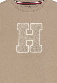 Maglione beige con logo "H" testurizzato in crema, dotato di scollo a giro e dettaglio con bottone sulla spalla. Tessuto morbido.